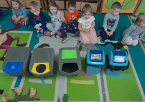 Dzieci siedzą na dywanie w kole, na środku którego znajdują się pojemniki do segregacji odpadów
