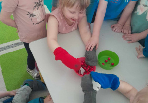 Chłopiec i dziewczynka próbują przenieść z jednego do drugiego pojemnika plastikowe wisienki za pomocą szczypiec mając na rękach skarpetki