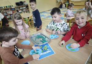 Dzieci siedzące przy stoliku wyklejają zieloną plasteliną niebieski kółka z paieru
